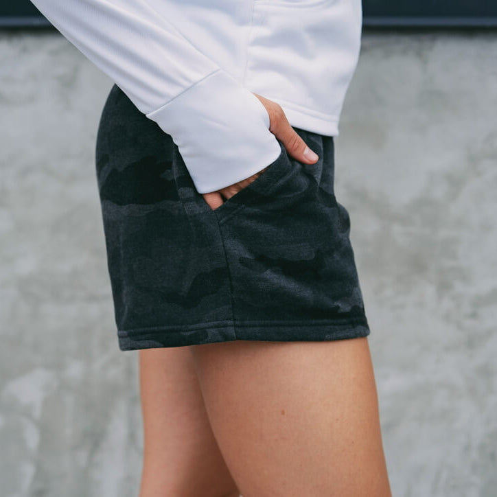 Person wearing a white long-sleeve shirt and black shorts against a gray concrete wall.