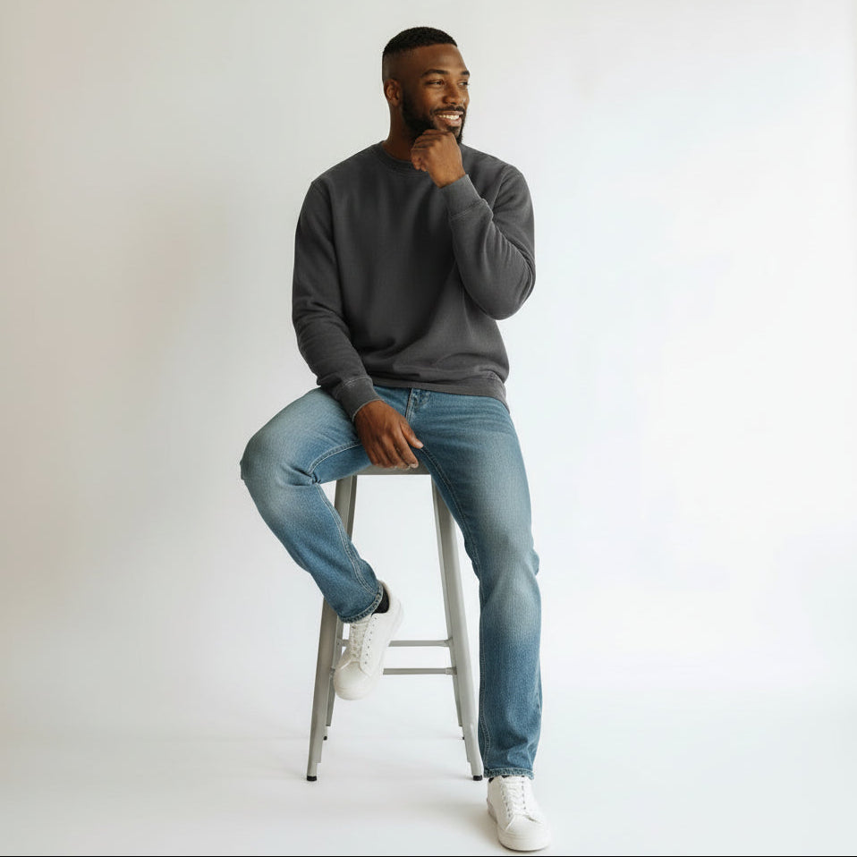 Man sitting on a stool wearing a gray sweater, blue jeans, and white sneakers against a plain background